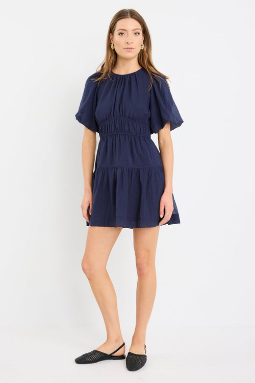 Woman wearing a navy mini dress with puff sleeves, elastic waist, and tie-back detail, styled with black flats.