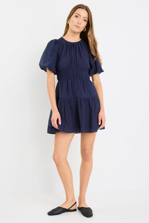Woman wearing a navy mini dress with puff sleeves, elastic waist, and tie-back detail, styled with black flats.