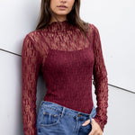 Woman wearing a burgundy sheer top and blue jeans against a white wall.
