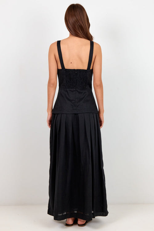 A woman viewed from the back wearing a black sleeveless maxi dress with a smocked bodice and flowing skirt, standing against a plain white studio background.