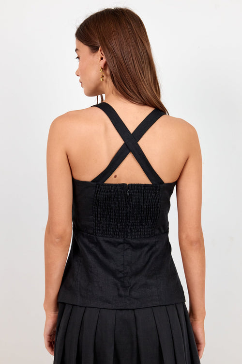 A woman shown from the back wearing a black sleeveless dress with crisscross shoulder straps, a smocked bodice, and a pleated skirt against a plain light background.