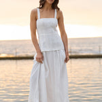 A woman wearing a white sleeveless linen top and matching white maxi skirt stands on a concrete pier with the ocean and sunset in the background.