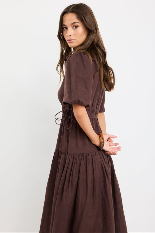 Woman wearing a chocolate brown linen midi dress with puff sleeves, tie waist detail, and tiered skirt, styled with black strappy sandals.