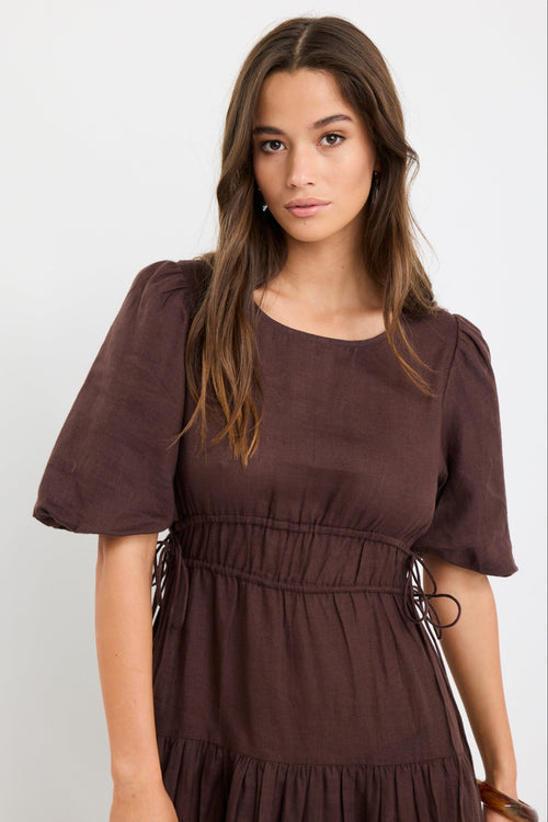 Woman wearing a chocolate brown linen midi dress with puff sleeves, tie waist detail, and tiered skirt, styled with black strappy sandals.