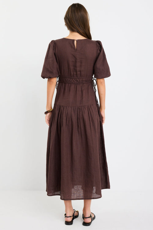 Woman wearing a chocolate brown linen midi dress with puff sleeves, tie waist detail, and tiered skirt, styled with black strappy sandals.