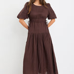 Woman wearing a chocolate brown linen midi dress with puff sleeves, tie waist detail, and tiered skirt, styled with black strappy sandals.