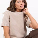 Woman wearing a beige sweater and brown pants against a white background