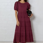 Woman wearing a burgundy dress holding a black handbag against a white brick wall.