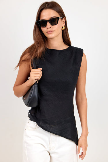 A woman modeling a sleeveless black top with white pants, black sunglasses, and a small black shoulder bag against a plain light background.