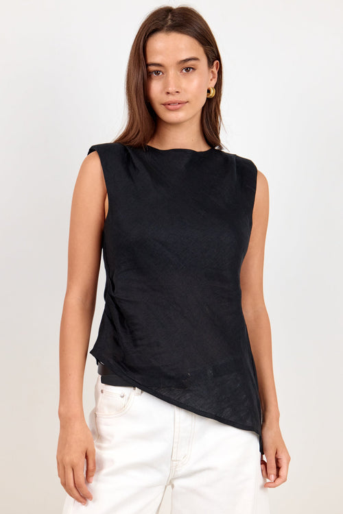 A woman modeling a sleeveless black top with a relaxed, slightly asymmetrical hem, paired with white pants, against a plain light gray studio background.