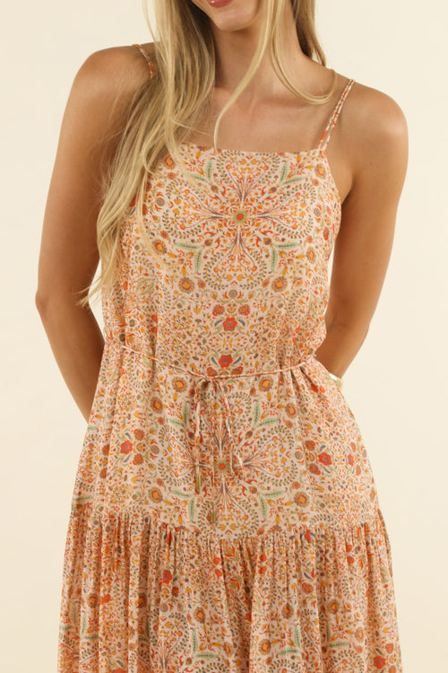 A woman wearing a sleeveless tiered dress with a dense orange, peach, and beige floral mandala pattern against a solid cream background.