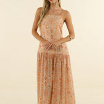 A woman modeling a sleeveless, tiered maxi dress with a small orange and green floral print on a light peach-beige base, set against a plain cream studio background.