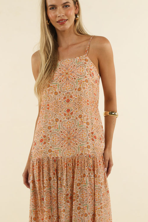 A woman wearing a sleeveless tiered maxi dress with a warm-toned bohemian floral and geometric pattern in orange, beige, and sage green, set against a solid cream background.