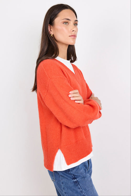 Woman wearing an orange sweater over a white shirt with blue jeans on a plain background