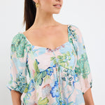 woman wearing a flowing floral puff sleeve dress with elastic waist and pastel blue and green print, styled with tan sandals.