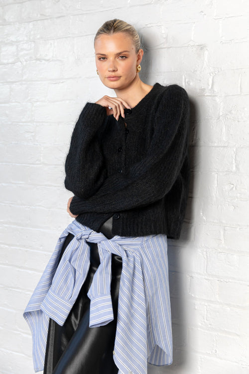 Woman wearing a black sweater and striped shirt against a white brick wall