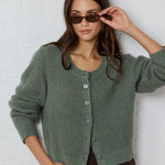 Woman wearing a green knitted cardigan and sunglasses against a white brick wall.
