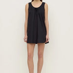 A woman modeling a sleeveless black tunic dress with a tie-neck detail, paired with black flat shoes, against a plain off-white studio background.