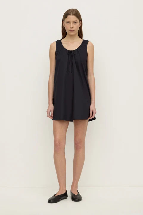 A woman modeling a sleeveless black tunic dress with a tie-neck detail, paired with black flat shoes, against a plain off-white studio background.