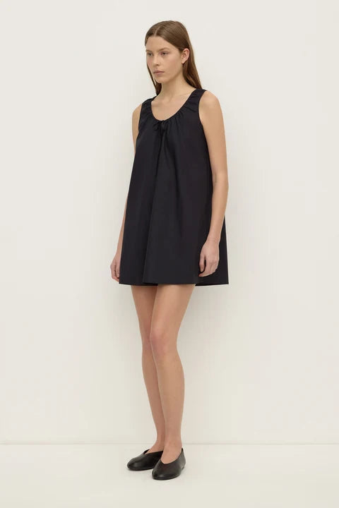 A woman modeling a sleeveless black mini dress with a gathered neckline and flared skirt, paired with black flats, against a plain off-white studio background.