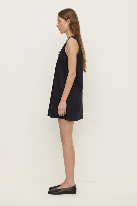 A woman stands in profile wearing a sleeveless black mini dress and black flats against a plain off-white studio background.