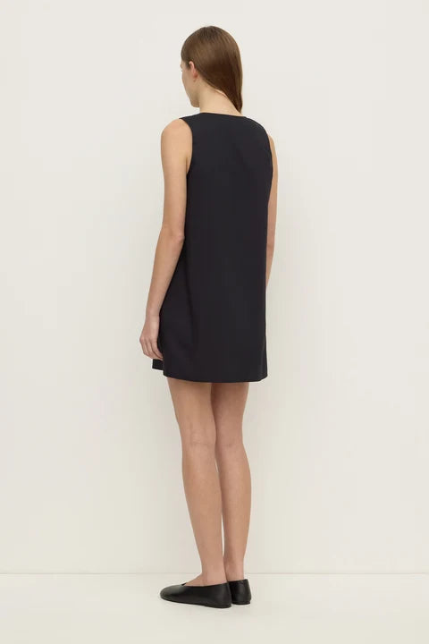 A woman stands with her back to the camera wearing a sleeveless black shift dress and black flats against a plain white studio background.