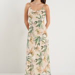 Woman wearing a bias-cut midi dress with a tropical palm print, strappy sleeves, and a softly draped silhouette, styled with neutral sandals.