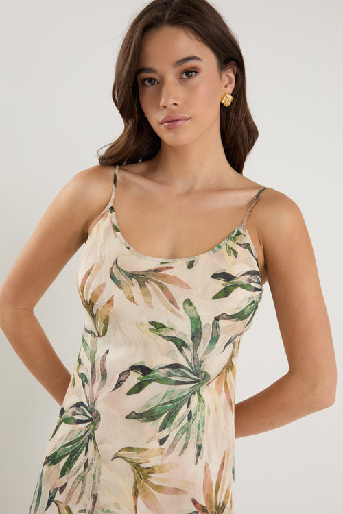 Woman wearing a bias-cut midi dress with a tropical palm print, strappy sleeves, and a softly draped silhouette, styled with neutral sandals.