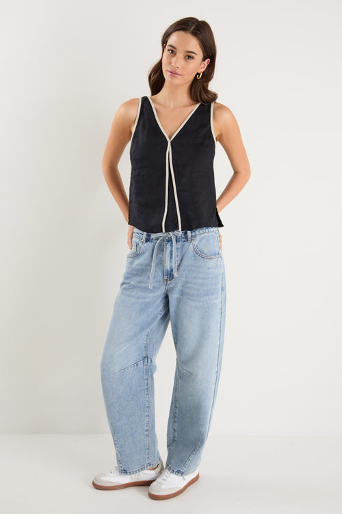 Woman wearing a sleeveless black linen top with contrast beige trim, styled with relaxed mid-blue denim jeans.