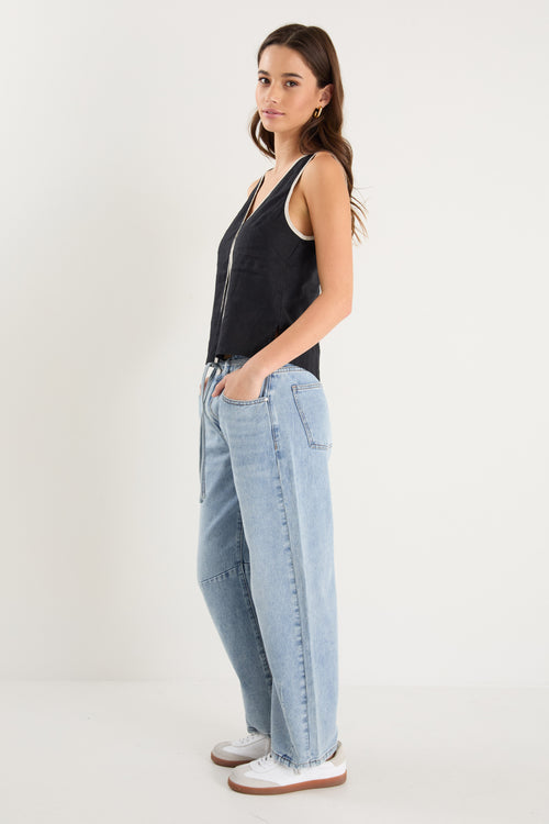 Woman wearing a sleeveless black linen top with contrast beige trim, styled with relaxed mid-blue denim jeans.
