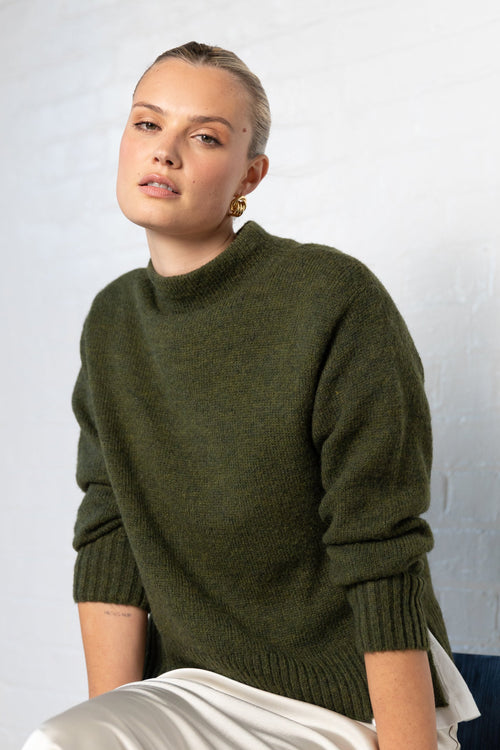 Woman wearing a green sweater sitting on a chair against a white wall