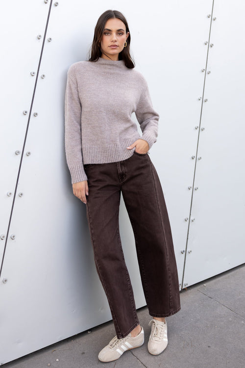 Woman in a gray sweater and brown pants standing against a white wall.