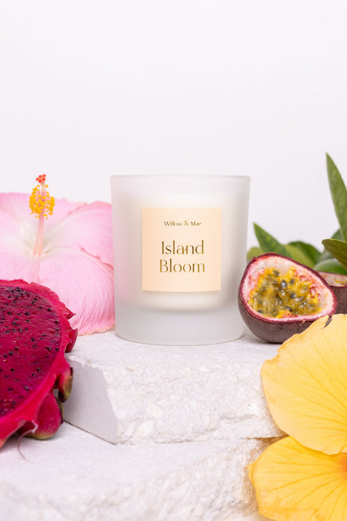 Candle labeled 'Island Bloom' surrounded by tropical fruits and flowers on a white surface.