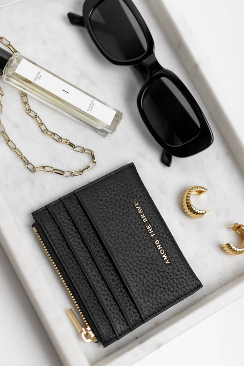 Black cardholder, sunglasses, perfume bottle, necklace, earrings on a white surface