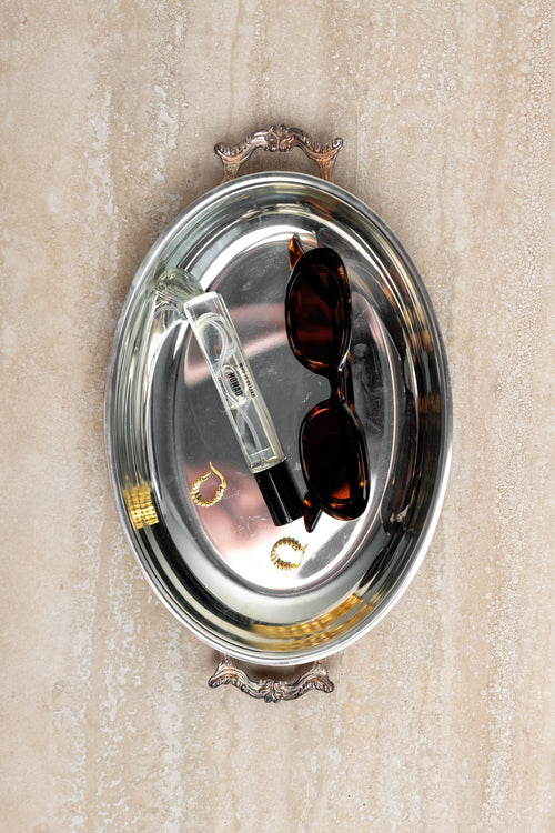 Silver tray with sunglasses, perfume bottle, and jewelry on a beige background