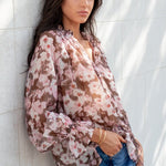 Woman leaning against a white wall wearing a floral blouse and blue jeans.