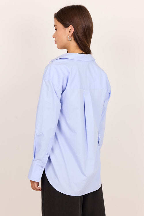 A woman shown from the back wearing a light blue long-sleeve button-up shirt and black shorts against a plain beige studio background.