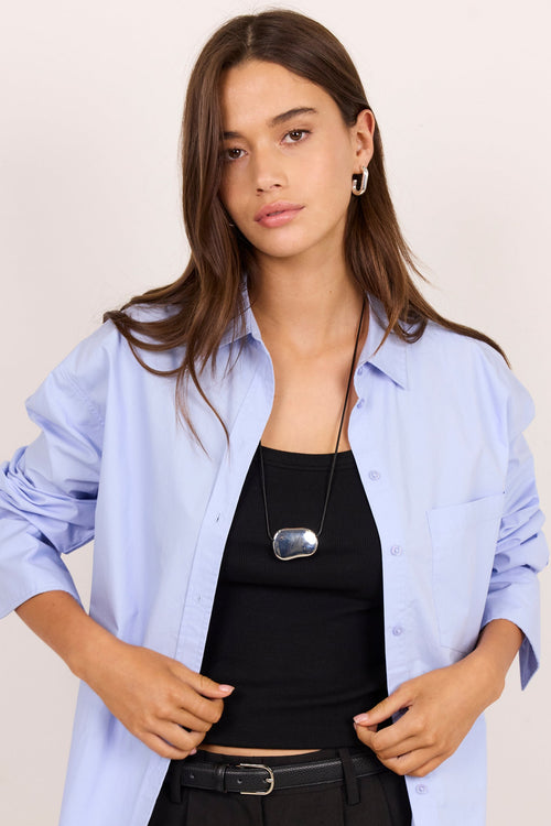 A woman modeling a light blue button-down shirt worn open over a black tank top and black trousers, photographed against a plain light beige studio background.