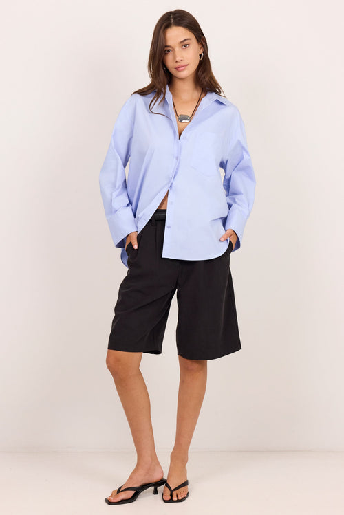 A woman modeling a light blue button-down shirt and black shorts against a plain off-white studio background.