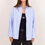 A woman modeling an oversized light blue button-up shirt over a black tank top and black shorts, photographed against a plain light beige studio background.