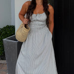 Woman in a light gray dress holding a straw bag outside a modern house.