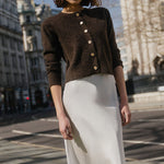 Woman in a brown cardigan and white skirt standing on a city street with buildings in the background