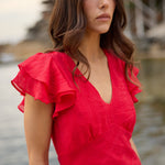 A woman wearing a bright red wrap-style dress with ruffled short sleeves, standing outdoors near water with a blurred waterfront setting in the background.