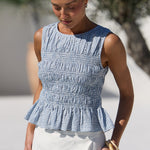A woman wearing a sleeveless smocked peplum top in a light blue and white gingham pattern with a white skirt, standing outdoors near a poolside lounge chair.