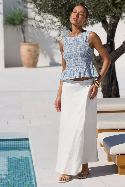 A woman stands by a poolside patio wearing a light blue smocked sleeveless top and a long white skirt. Sun loungers, a tree, and white walls are visible in the background.