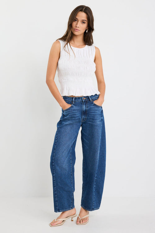 Woman wearing an ivory linen sleeveless top with a shirred bodice and ruffle hem, paired with dark blue jeans.