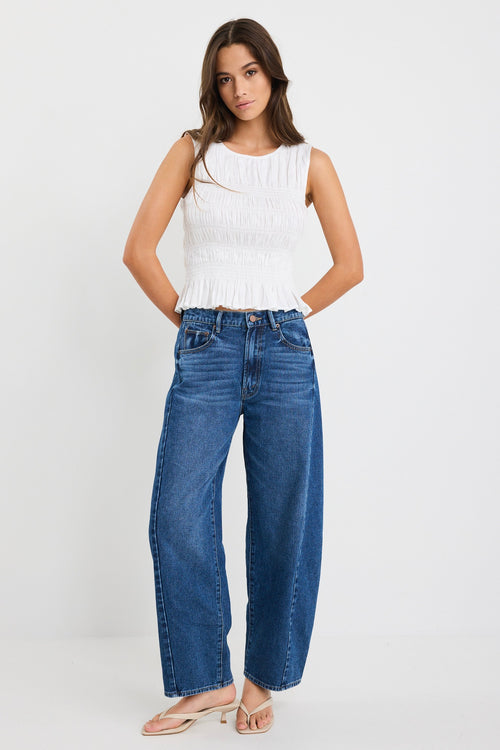 Woman wearing an ivory linen sleeveless top with a shirred bodice and ruffle hem, paired with dark blue jeans.