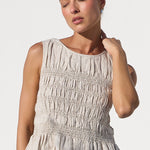 A woman modeling a sleeveless beige smocked peplum top and wide-leg light blue jeans, standing in a bright white architectural setting with arches.