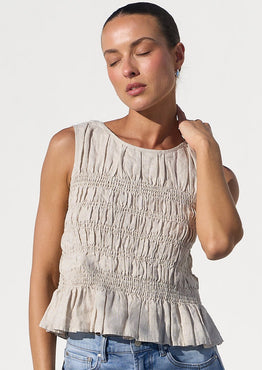 A woman modeling a sleeveless beige smocked peplum top and wide-leg light blue jeans, standing in a bright white architectural setting with arches.