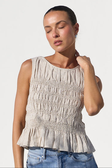 A woman modeling a sleeveless beige smocked peplum top and wide-leg light blue jeans, standing in a bright white architectural setting with arches.
